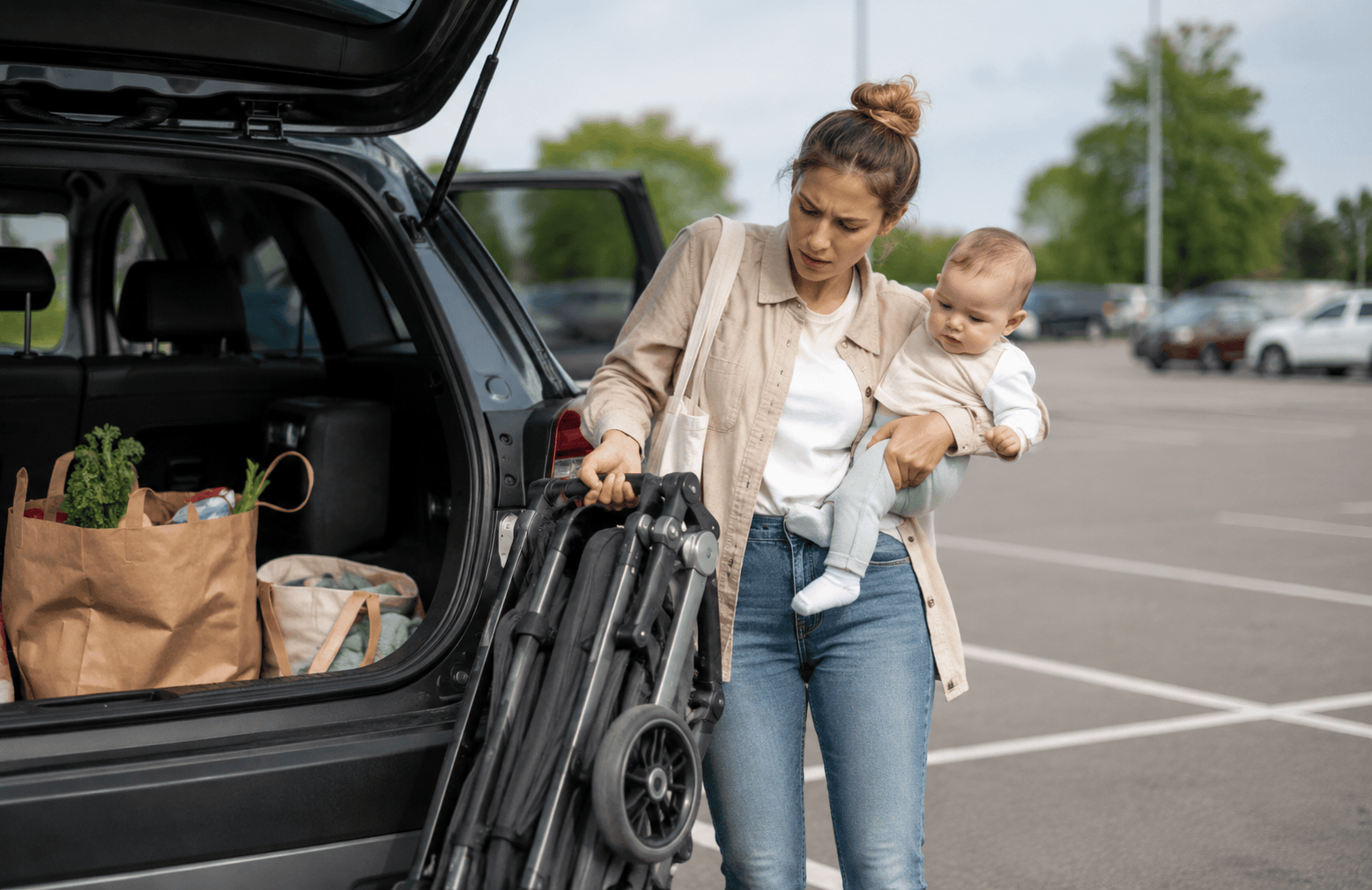 Anyuka babával és összecsukott babakocsival az autó mellett egy parkolóban.